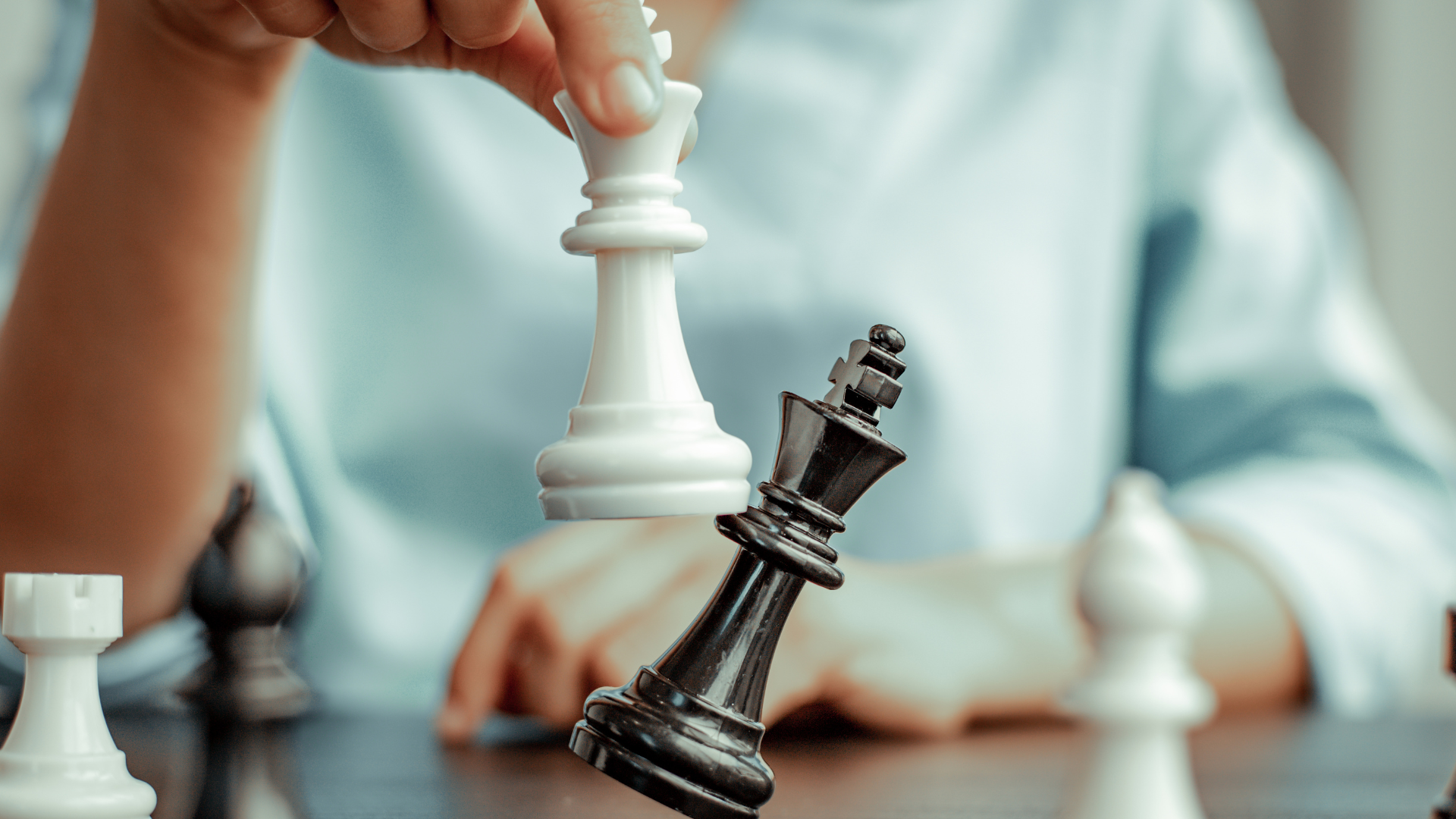 Person's hand holds a white queen chess piece to knock over a black king chess piece. Other chess pieces surrounding.