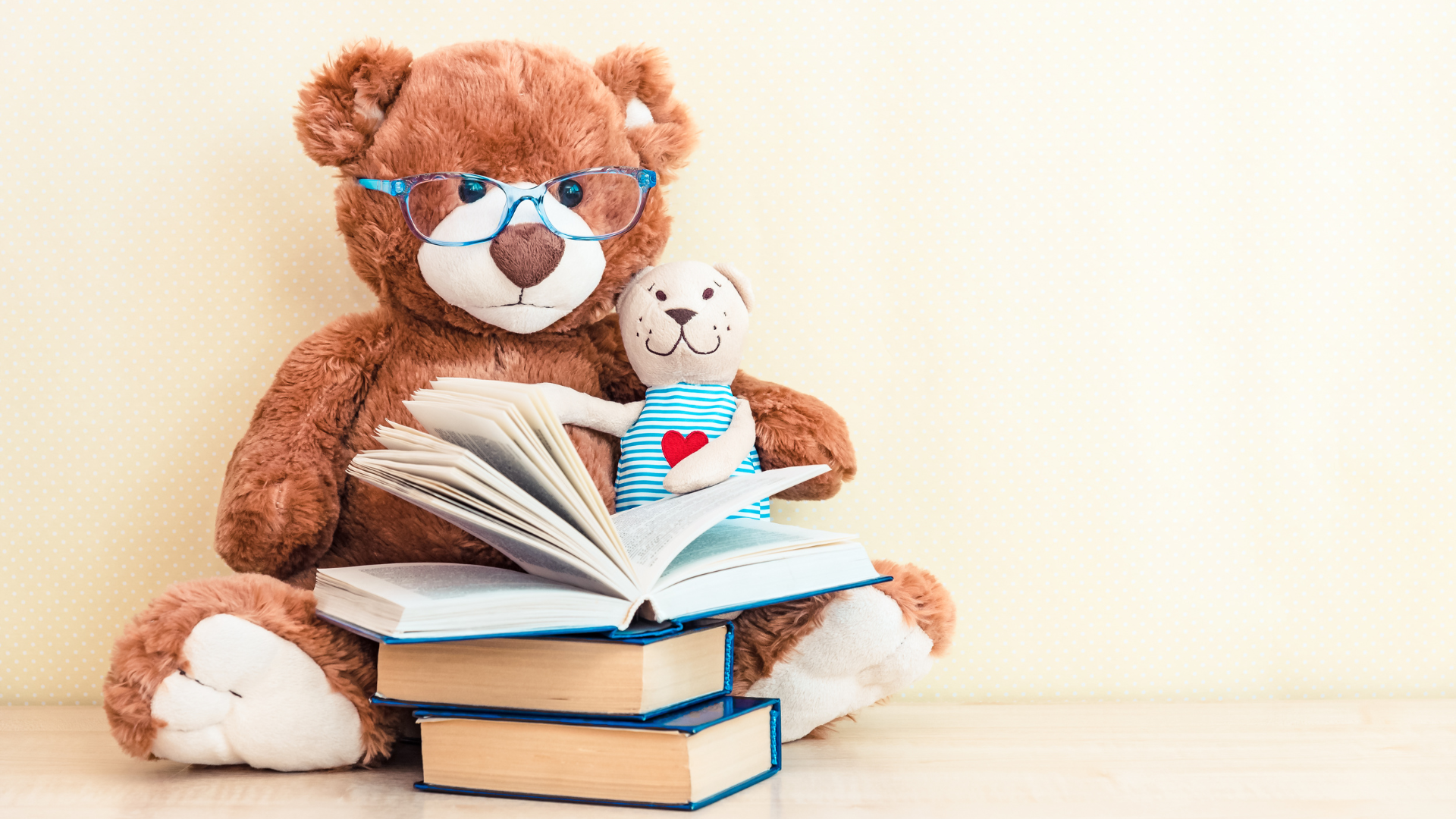 A brown stuffed bear wearing glasses sits with a stack of books, the one on top opened, and a smaller cream-colored stuffed bear on its lap.