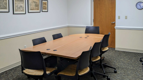 Image of the Conference Room. Wooden tables arranged in the center of the room, surrounded by rolling chairs. Door and clock on the wall on the far end, and four visible framed photographs on the wall to the left.