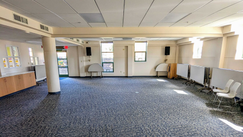 Image of the Meeting Room. Blue abstract patterned carpet, five visible windows, a counter with wooden cabinets and a pillar to the left. Tables and chairs are put away and stacked to the right.