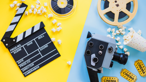 Yellow and blue background split diagonally in the middle. On the left is a clapperboard, scattered popcorn, and a film reel. On the right is another film reel, popcorn, a video camera, and three tickets that say "cinema"
