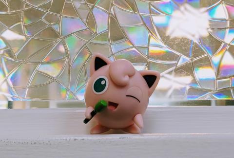 A figurine of the Pokemon Jigglypuff (small pink ball with triangle cat-like ears a curl on its forehead and small rounded arms and legs) winking one eye and holding a microphone up to its mouth stands in front of a swirly iridescent background