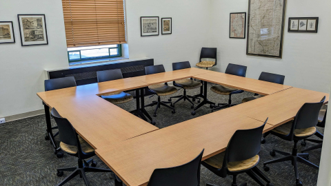 Image of the Presentation Room with six wooden rectangular tables arranged in a square with rolling chairs placed around them. A window and six framed photographs are on the walls.