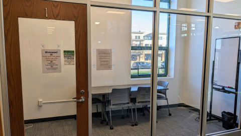Looking into the door and windows, there is a rectangular table surrounded by four chairs in the quiet study room 1. There is also a window to the park and a dry erase board.