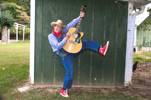 Man in a cowboy hat, overalls, neckerchief, and red converse high-tops holds a guitar up and kicks his left left up in the air