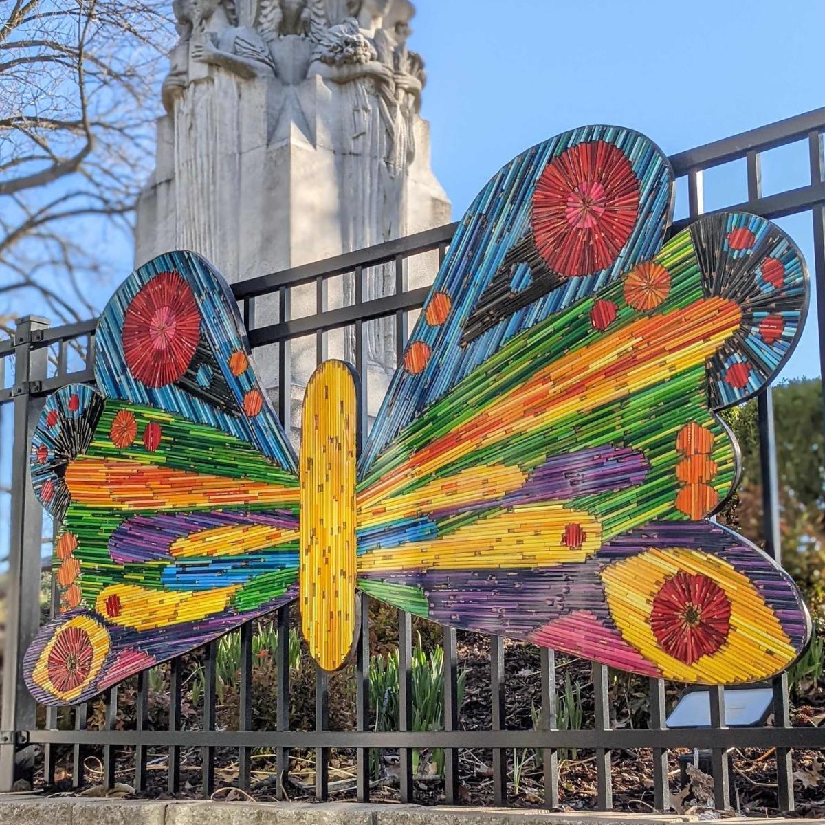 Art piece of a butterfly made from colored pencils hanging from a black metal fence.