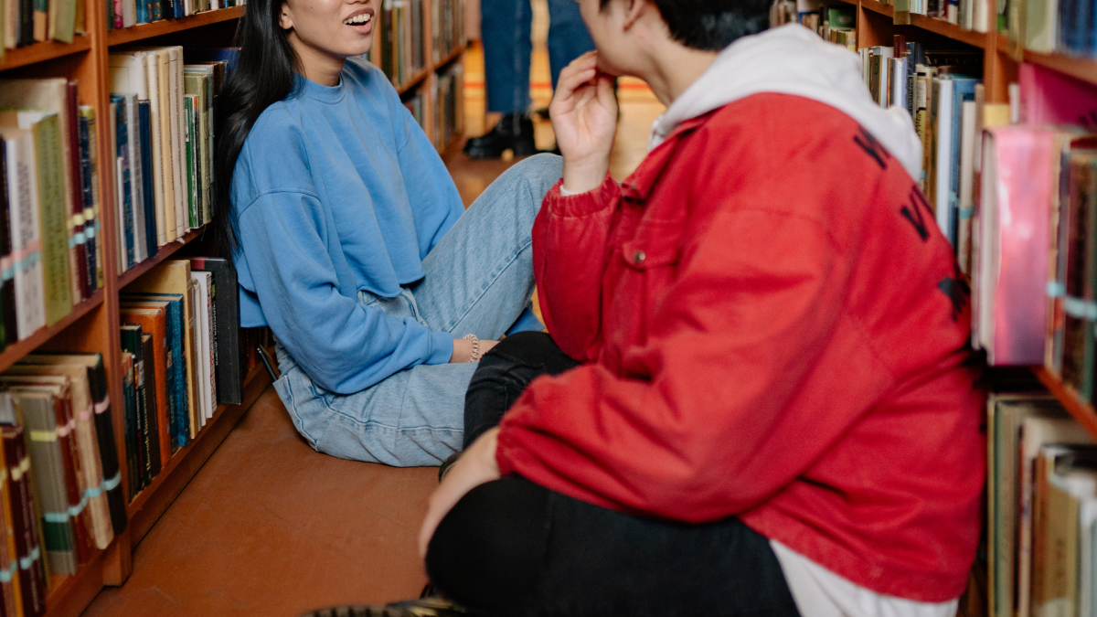 Two young people sit chatting between library shelves